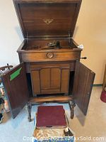 Front view of open wooden console cabinet phonograph with top lid and front doors open, tone arm and grille below, red case on floor