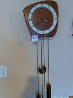 Front view of the triangular wooden clock with white and bronze circular face, gold-tone numerals and hands, and three hanging chains supporting pendulum and weight.