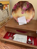Wooden box with red fabric lining displaying Community Plate flatware and hand-painted porcelain plate with pink flowers on gradient background.