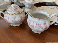 Close-up of sugar bowl and creamer showing pink rose pattern with gold trim on white porcelain surface, scalloped edges visible