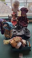 Five vintage dolls arranged on table with pink baby doll cradle, cast iron miniature stove, and box of doll clothes.