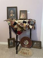 Full view of half moon table with crazy quilt on top, framed artworks behind and around the table, decorative plate in front, small hand knotted floral rug on floor.