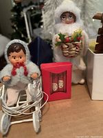 Close-up of vintage-style Christmas dolls and miniature white pram with tangled cords, plus a tiny boxed pair of Christmas crackers