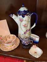 Overall lot showing the white floral cocoa jug with blue accents, pink and gold Paragon teacup and saucer, small cobalt blue teapot figurine, small floral ashtray with pink rose, and small gradient blue ceramic container.