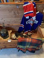 Open wooden chest showing Old Irving Oil Co. jersey draped inside along with black miner's helmet and a plaid wool blanket partially hanging over the edge.