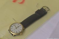 Photo of a men's wristwatch featuring a white dial with black Roman numerals, gold-tone case, and black leather strap with silver buckle.