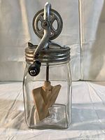 Front angled view of the butter churn showing glass jar, wooden paddle, and metal crank with handle.