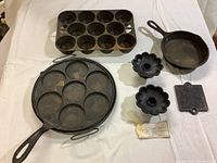 Overview of all seven cast iron pieces on white cloth showing muffin pan, plett pan, small skillet, large skillet/hot plate, flower-shaped candle holders, and trivet.