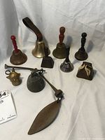 Full set of 9 vintage bells arranged on cloth background showing variety of sizes and shapes including hand bells, cowbells, chime bell and clown bell