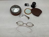 Photo showing all six vintage optical items on white background: round wireframe glasses, large magnifying glass, small magnifying glass with case, black jeweler's loupe, and metal folding magnifier.