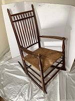 Full view of the older oak rocking chair showing the vertical slatted back, finial posts, wooden armrests, and woven rattan seat.