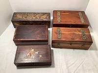 Six wooden boxes of varying sizes and styles arranged on white surface. Top three include a long narrow decorative box, two medium rectangular wooden boxes with brass hardware. Bottom three are smaller decorative wood boxes with inlaid or painted motifs.
