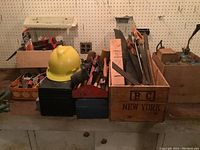 Overview of workbench with various tools in wooden crates and boxes, showing saws, hammers, mallets, and a yellow hard hat.