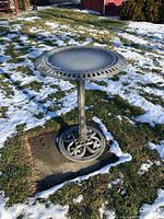 Bird bath standing on grass with patches of snow, showing full height and ornate base.