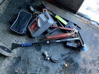 Overview photo showing multiple car jacks, a black bag, a red hydraulic jack, oil can, scraper, and assorted automotive tools and parts on a concrete floor.