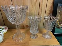 Wide view of all three crystal pieces on wooden surface: a tall vase with scalloped edge, a shorter vase with detailed cut glass, and a stemmed bowl with fan patterns.