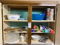 Photo of cupboard shelf showing CorningWare casserole dish with cornflower pattern, plastic blue mixing bowl, cake tray, plates, kitchen wrap boxes, and Pringles cans