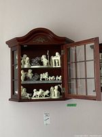 Faux wood wall mounted display cabinet with glass door left open showing three shelves with Snowbabies figurines inside