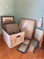 Photo showing a box filled with multiple picture frames of various sizes leaning on the floor with some laid out in front.