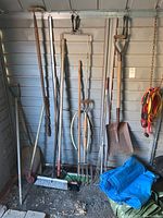 Photo of various gardening tools leaning against a wall including shovels, rakes, pitchfork, broom, small brush and blue tarp on the floor.