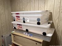 Four clear plastic under bed storage boxes stacked, three with wheels and one without, on a wooden surface against wall paneling.