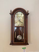 Front view of wooden wall clock with brass face, Roman numerals, and decorative elements.