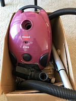 Front and side view of pink Bissell Zing canister vacuum in cardboard box with hose, metal wand and various attachments visible.