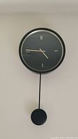 Black framed wall clock with pendulum hanging on wall, minimalist dial with partial hour marks.