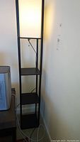 Full view of the tall floor lamp in a corner with two square shelves, black metal frame and illuminated fabric shade.