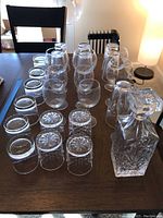 Photo showing a crystal decanter with stopper and assorted various sized glasses including tumbler glasses with patterned bases on a dark wood table.