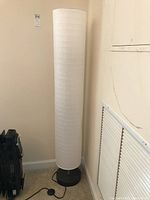 Tall white cylindrical paper floor lamp standing next to a wall, showing base and cord with foot switch.