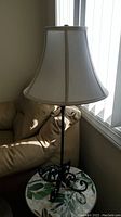 Full view of metal base table lamp with cream fabric bell-shaped lampshade, positioned next to a couch and window