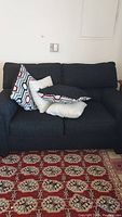 Front angled view of loveseat with multiple decorative cushions arranged on seating and backrest. Dark fabric upholstery visible. Loveseat positioned on patterned red and beige carpet.