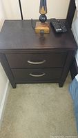 Full view of one dark wood nightstand next to a bed with remote controls and lamp on top, showing two drawers and short legs.
