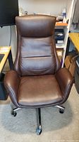 Front view of the brown leather high back office chair showing padded seat, backrest, headrest, and arms.