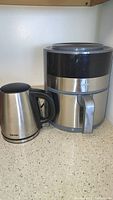 Photo showing Aroma electric kettle beside Crux air fryer on countertop, both stainless steel with black accents.