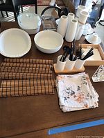 Overview of ceramic dishware, Corning casserole with lid, ceramic vases, cutlery holder with black utensils, napkin holders, bamboo placemats, and cloth placemats with floral design on a wooden table.