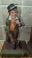 Front view of The Artful Dodger figurine wearing green coat, yellow vest, polka-dotted bow tie, black top hat, standing on book-shaped base
