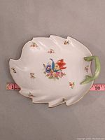 Top view of a leaf-shaped porcelain dish with floral pattern, green handle, and gold trim with measuring tape showing approx. 9.5 inches length.