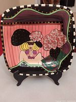 Front view of square platter showing colorful illustration of woman with black hat and pink floral decoration.