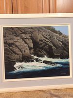 Full view of framed painting showing rocky shoreline, waves and forest background.