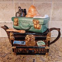 Front view of the teapot showing the mottled black and brown trunk base with gold accents, handle and spout, and the green suitcase lid adorned with a safari hat, binoculars, tea kettle, and travel stickers.