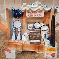 Close-up showing the miniature kitchenware decorations on the teapot base and lid, including tiny plates, cups, bowls, a grater, and salt and pepper shakers, along with the 'Woodmanton China Co.' marking and branding on the front.