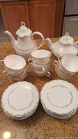 Photo shows stacked teacups and saucers with dessert plates in front, alongside the coffee pot and teapot at the back on the kitchen counter.
