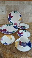 Full set of dinnerware with plates, bowls, cups, and serving pieces arranged on granite countertop, showing floral pattern on white china.