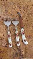 Three cutlery pieces with white bone china handles decorated with Royal Albert Old Country Roses floral pattern. Two serving forks and one cheese cutter with stainless steel tips.