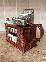 Side view of the large ceramic teapot resembling a wooden teashop counter with multicoloured tea canisters and a shiny vintage cash register on top.