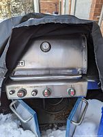 Front view of Weber Genesis stainless steel grill with three control knobs, lid thermometer, handle and torn cover