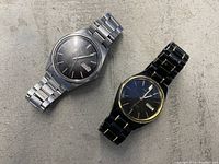 Photo shows two vintage Seiko watches: one silver stainless steel with black gradient dial, and one black with gold-tone accents. Both have day-date windows.