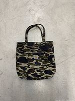Front view of Carhartt WIP camo tote bag showing full bag and straps laid flat on concrete floor.
